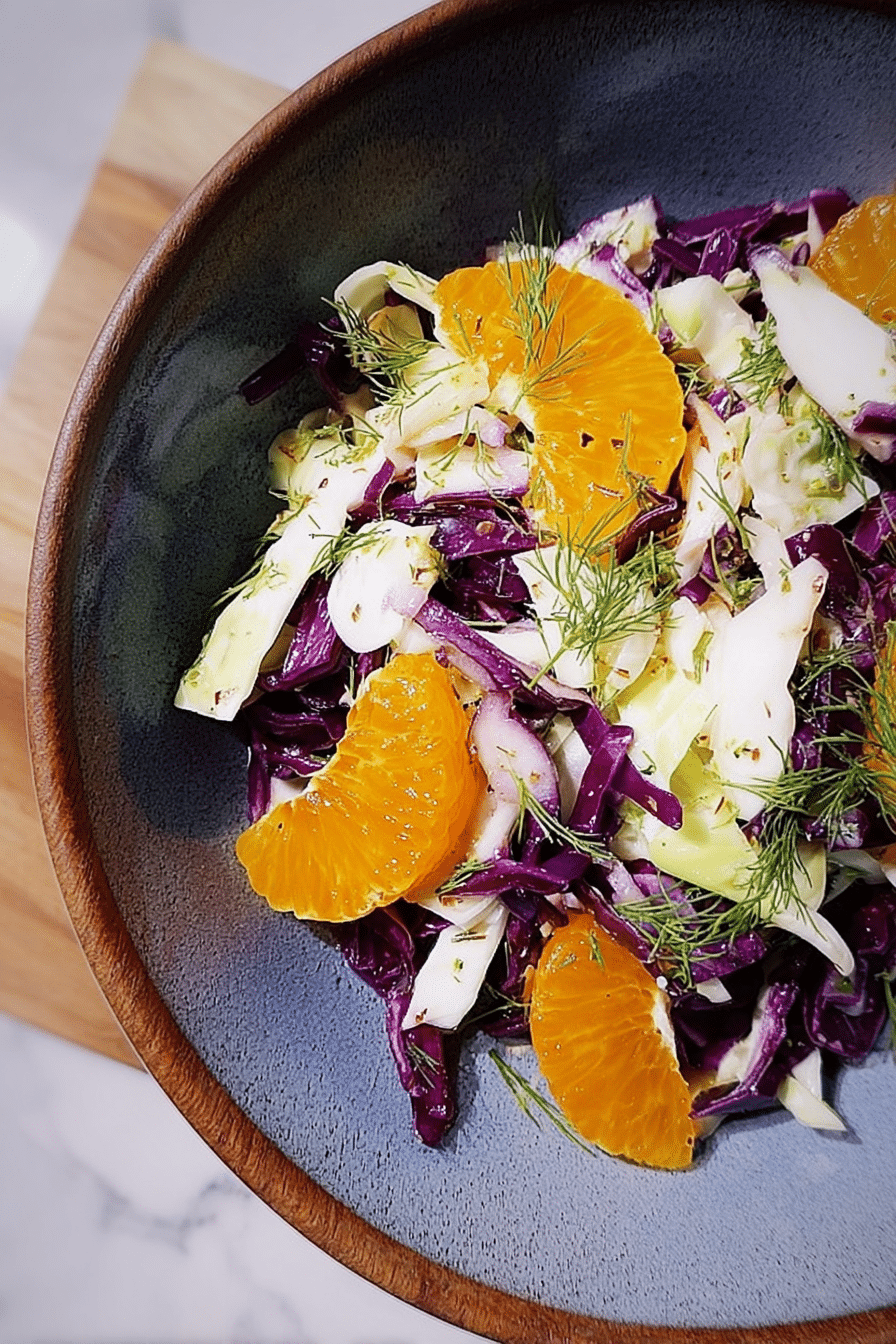 Bowl of cabbage, fennel, and citrus salad garnished with fennel fronds on a wooden table.