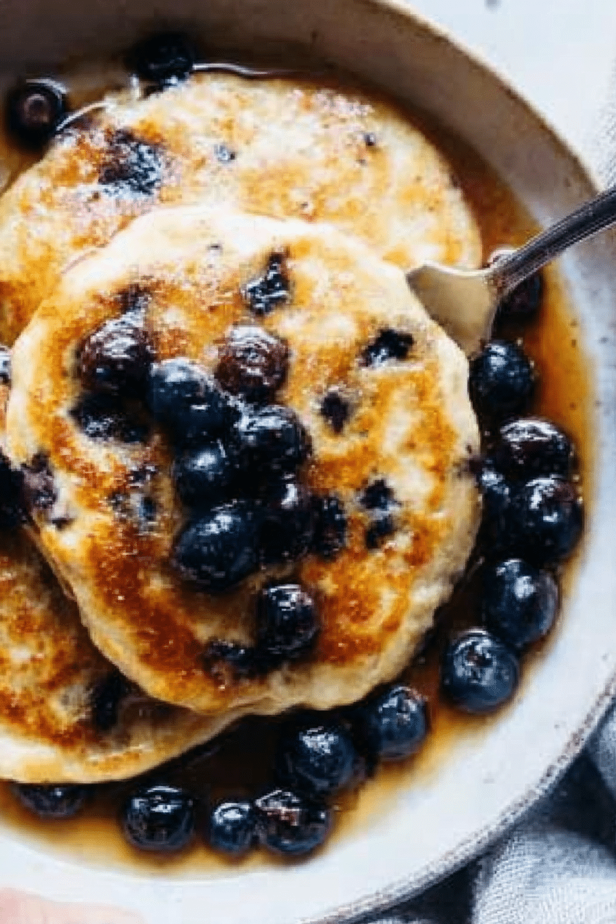 A delicious stack of fluffy blueberry pancakes with fresh blueberries and maple syrup on a wooden table.