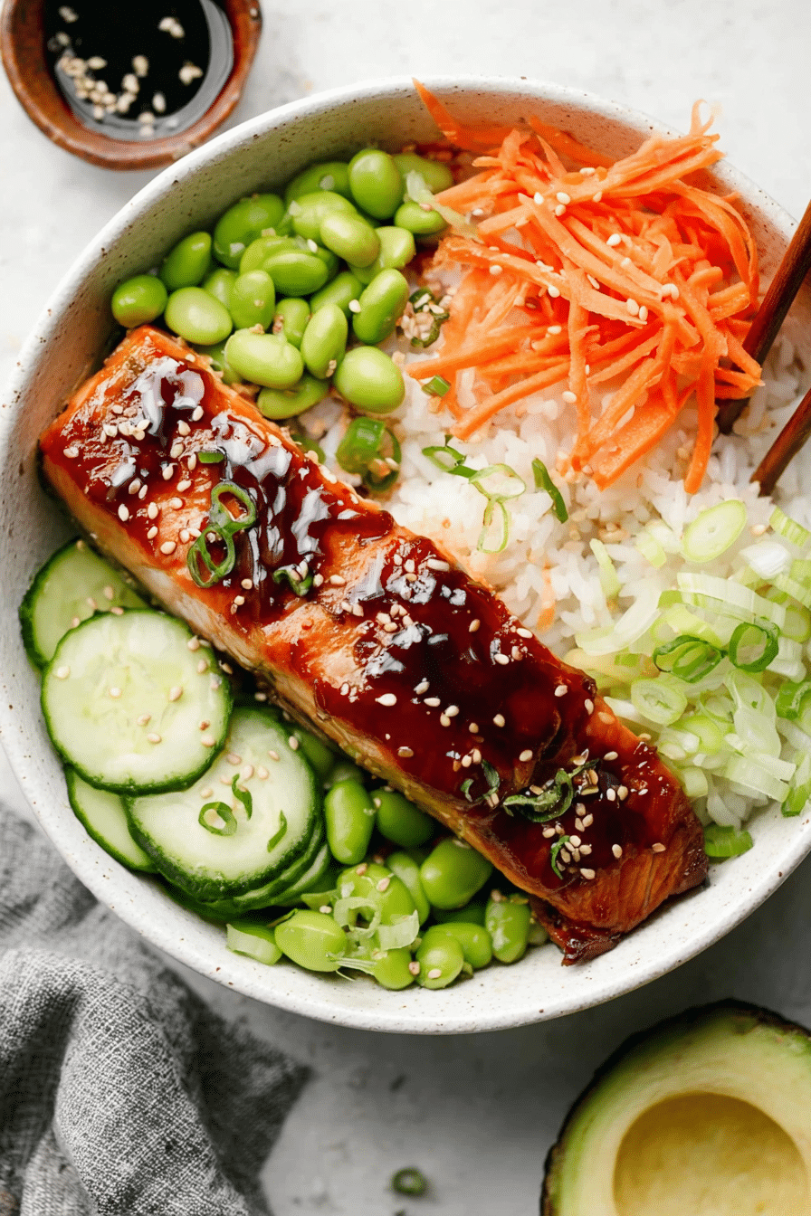 Delicious teriyaki salmon bowl with salmon fillet, brown rice, cucumbers, edamame, carrots, and avocado served in a shallow bowl.