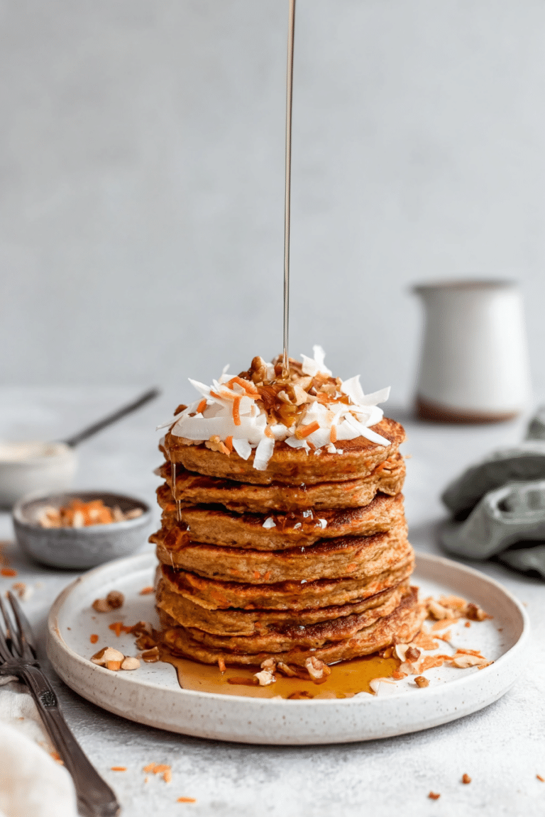 Fluffy carrot cake pancakes topped with cream cheese frosting, walnuts, and maple syrup, with carrots and spices in the background.