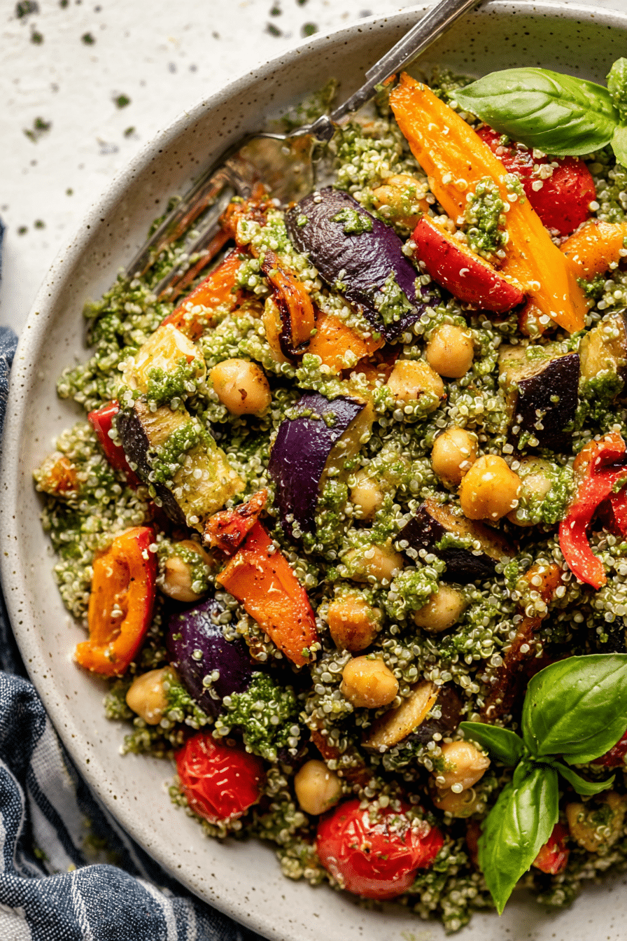 A colorful roasted veggie chickpea pesto quinoa salad with fresh basil, featuring roasted bell peppers, onions, and sweet potatoes, served in a bowl.