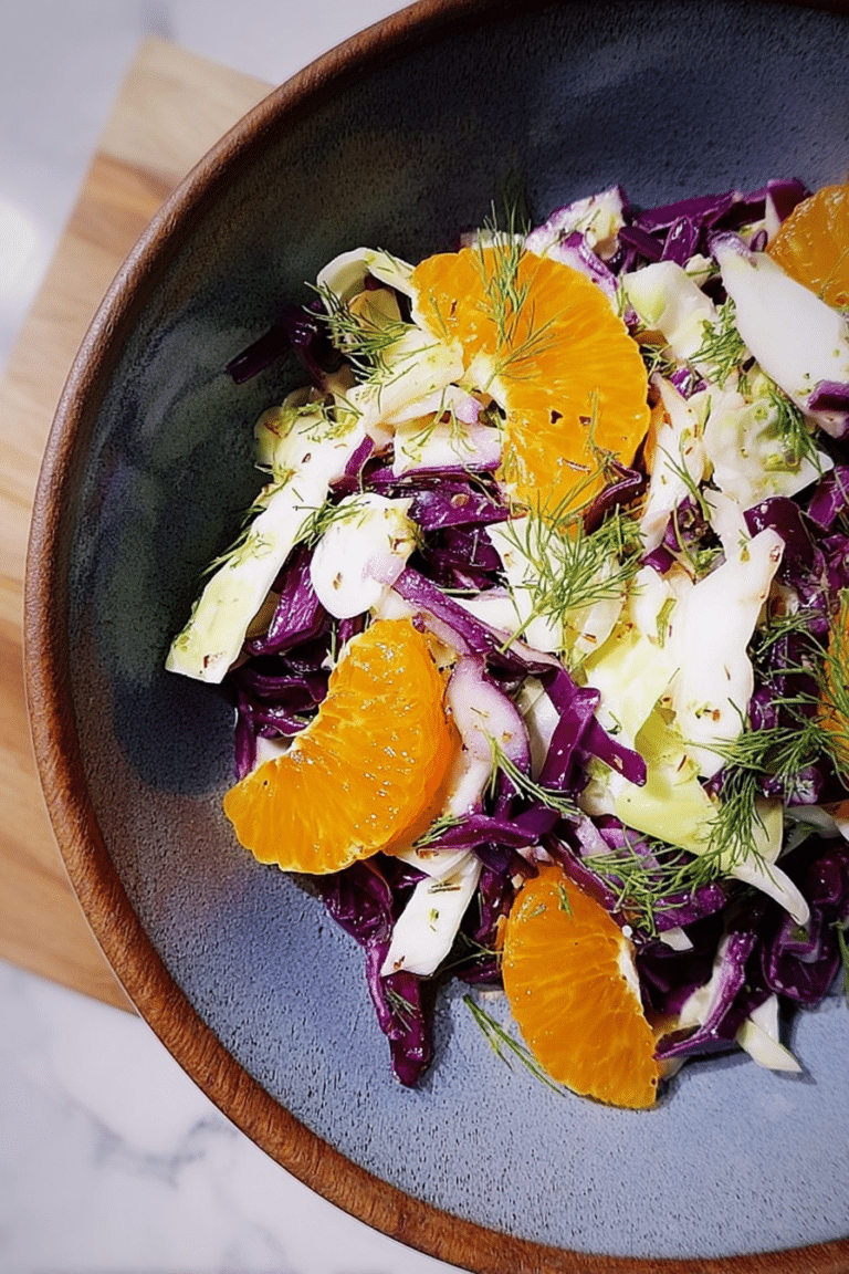 Bowl of cabbage, fennel, and citrus salad garnished with fennel fronds on a wooden table.